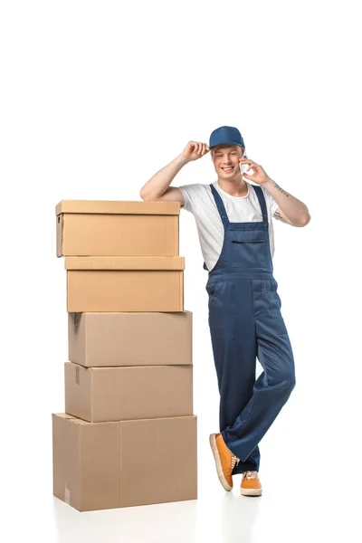 Guapo sonriente mover en uniforme con cajas de cartón hablando en teléfono inteligente aislado en blanco - foto de stock