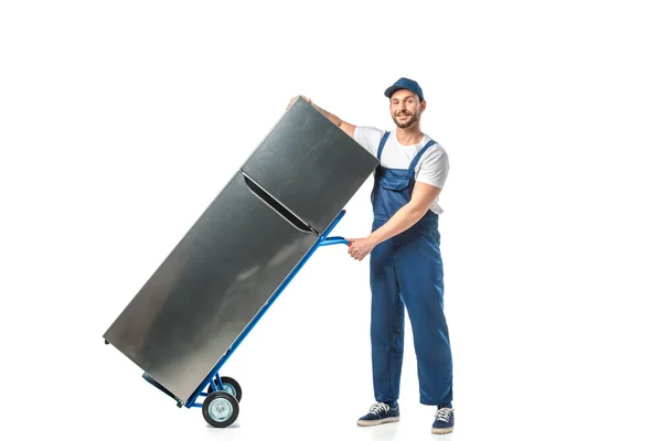 Handsome mover in uniform looking at camera and transporting refrigerator on hand truck isolated on white — Stock Photo