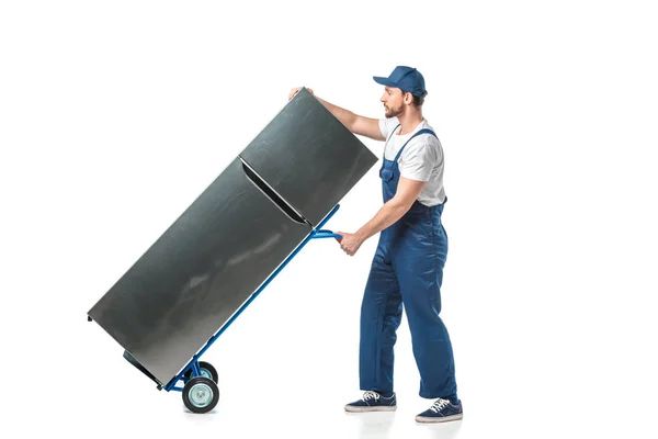 Handsome mover in uniform transporting refrigerator on hand truck isolated on white — Stock Photo