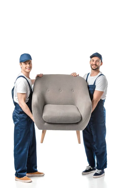 Два улыбающихся грузчика смотрят в камеру и перевозят серый кресло, изолированное на белом — стоковое фото