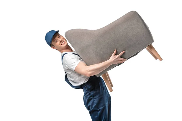 Handsome mover in uniform carrying grey armchair, smiling and looking at camera isolated on white — Stock Photo