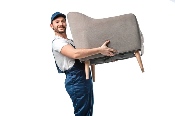 Bonito mover no uniforme olhando para a câmera enquanto carrega poltrona cinza isolado no branco — Fotografia de Stock