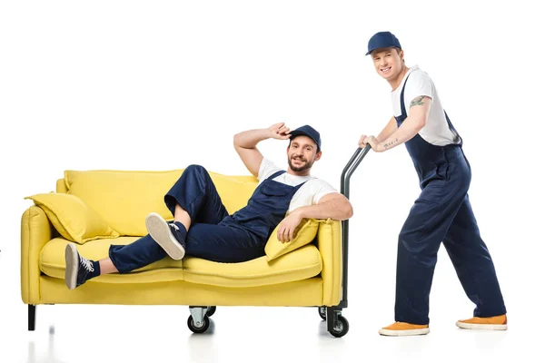 Mover in Uniform sitzt auf dem Sofa und blickt in die Kamera, während Kollege Handwagen auf weiß schiebt — Stockfoto