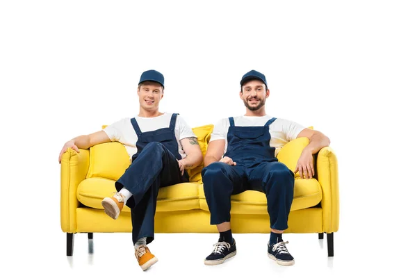 Due traslocatori sorridenti in uniforme seduti sul divano giallo e guardando la fotocamera isolata sul bianco — Foto stock