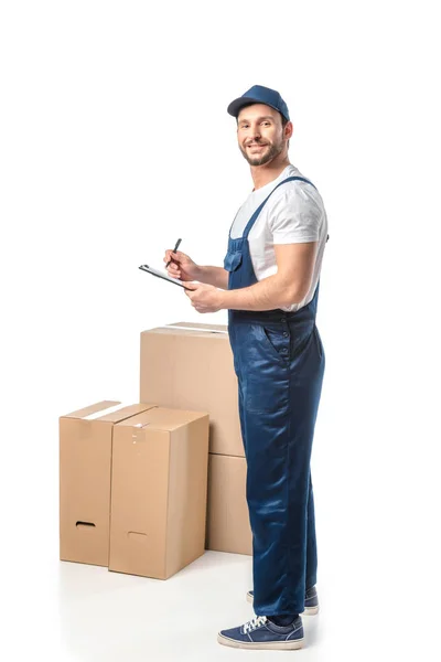 Déménageur souriant en uniforme écrit dans le presse-papiers près de boîtes en carton isolé sur blanc — Photo de stock