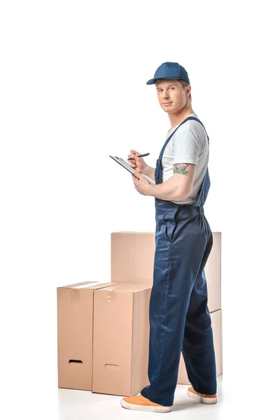 Mover in uniform writing in clipboard near cardboard boxes isolated on white — Stock Photo