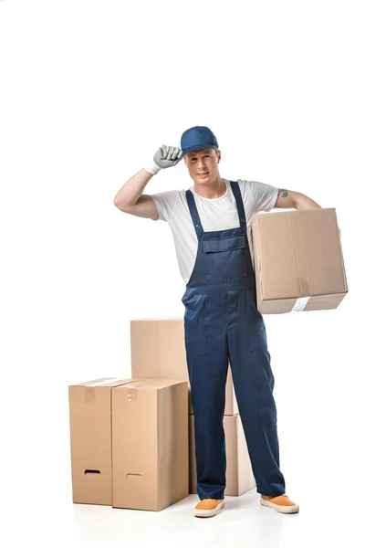 Handsome mover in uniform adjusting hat and looking at camera while carrying cardboard box isolated on white — Stock Photo