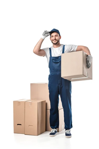 Guapo mover en uniforme ajustando sombrero y mirando a la cámara mientras lleva caja de cartón aislado en blanco - foto de stock