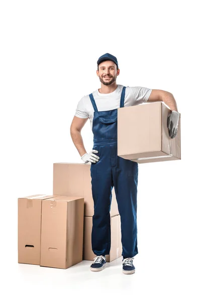 Bonito mover em uniforme transportando caixa de papelão, sorrindo e olhando para a câmera isolada no branco — Fotografia de Stock