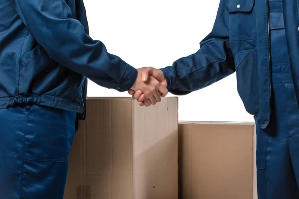 Vista recortada de dos transportistas en uniformes estrechando las manos cerca de cajas de cartón aisladas en blanco - foto de stock