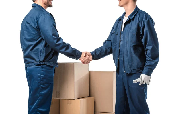 Vue recadrée de deux déménageurs en uniforme serrant la main près de boîtes en carton isolées sur blanc — Photo de stock