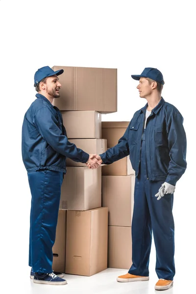 Two movers in uniform looking at each other and shaking hands near cardboard boxes on white — Stock Photo