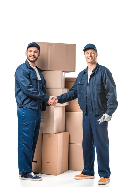 Two smiling movers in uniform looking at camera and shaking hands near cardboard boxes on white — Stock Photo