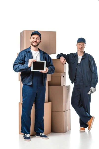 Two movers with cardboard boxes looking at camera and presenting digital tablet with blank screen on white — Stock Photo