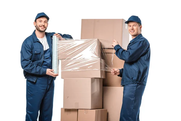 Deux déménageurs regardant la caméra et enveloppant des boîtes en carton avec rouleau de film étirable isolé sur blanc — Photo de stock