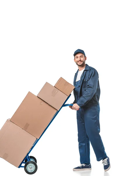 Smiling handsome mover in uniform transporting cardboard boxes on hand truck isolated on white — Stock Photo