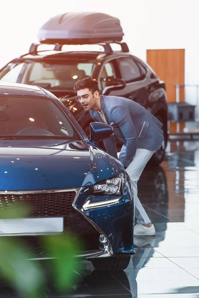 Homme élégant gai dans des lunettes debout près de l'automobile dans la salle d'exposition de voiture — Photo de stock