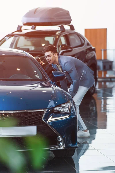 Cheerful stylish man in glasses standing near automobile in car showroom — Stock Photo