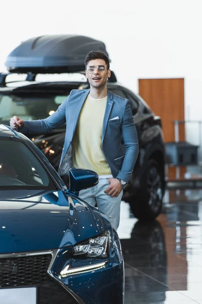 Hombre feliz en gafas de pie cerca de automóvil en sala de exposición de coches - foto de stock