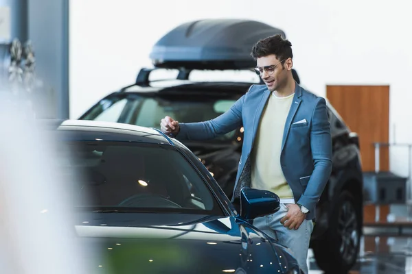 Selective focus of happy customer in glasses standing near automobile in car showroom — Stock Photo