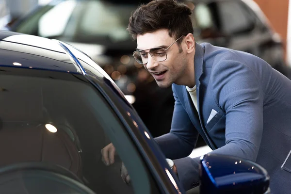 Happy customer in glasses standing near automobile in car showroom ac — Stock Photo