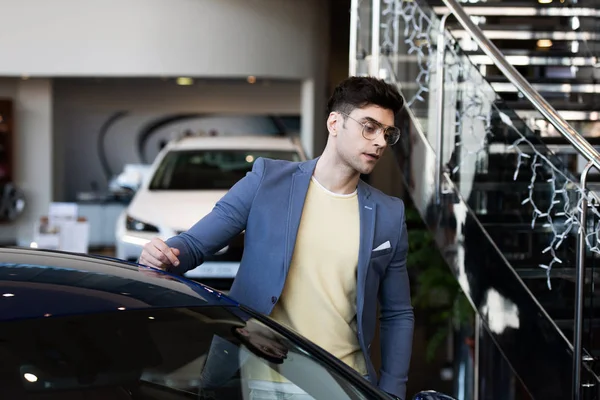 Handsome customer in glasses standing near automobile in car showroom — Stock Photo