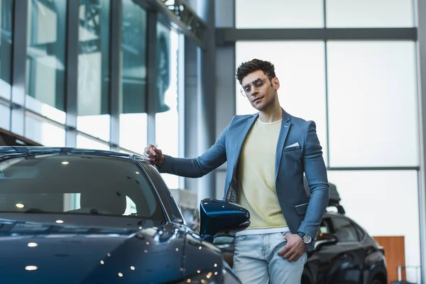 Cher homme dans des lunettes debout avec la main dans la poche près de l'automobile dans la salle d'exposition de voiture — Photo de stock