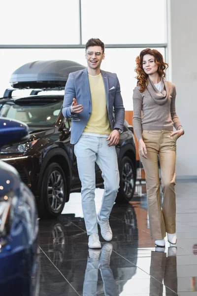 Hombre feliz en gafas apuntando con el dedo al automóvil cerca de la mujer rizada con la mano en el bolsillo - foto de stock