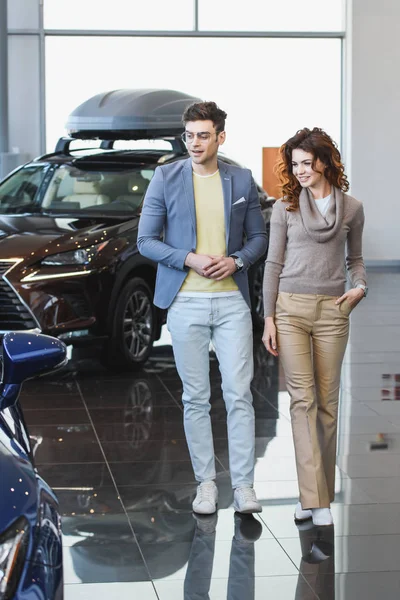 Hombre con estilo en gafas caminando en sala de exposición de coches cerca de mujer atractiva rizado con la mano en el bolsillo - foto de stock