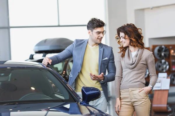 Hombre feliz en gafas apuntando con el dedo al automóvil cerca de la mujer rizada con la mano en el bolsillo - foto de stock