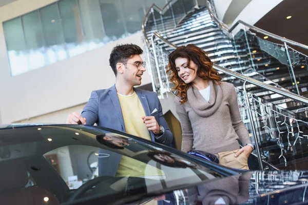 Stilvoller Mann mit Brille zeigt mit dem Finger auf Auto neben lockiger attraktiver Frau, die mit der Hand in der Tasche steht — Stockfoto