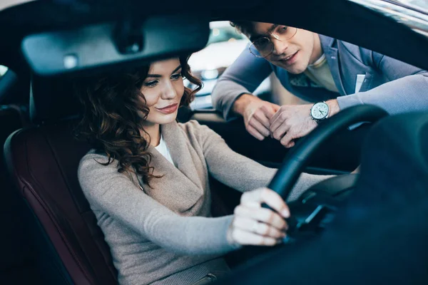Selektiver Fokus der schönen lockigen Frau, die im Auto sitzt und das Lenkrad neben einem fröhlichen Mann mit Brille hält — Stockfoto