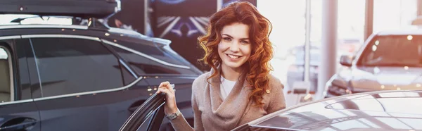 Panoramic shot of happy curly woman smiling while standing near vehicle in car showroom — Stock Photo