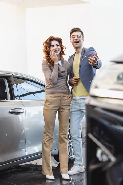 Foco seletivo de mulher encaracolada alegre de pé com o homem feliz apontando com o dedo para o carro — Fotografia de Stock