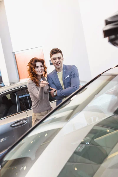Избирательный фокус веселой кудрявой женщины, указывающей пальцем на автомобиль рядом с красивым мужчиной — стоковое фото