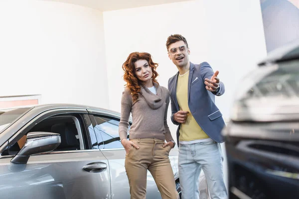 Selective focus of curly woman standing with hand in pocket near man pointing with finger at car — Stock Photo