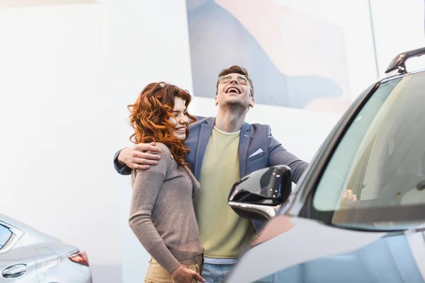 Foco seletivo de homem feliz abraçando mulher encaracolado no showroom carro — Fotografia de Stock