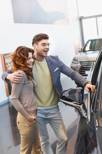 Feliz hombre abrazando rizado atractiva mujer de pie con la mano en el bolsillo en la sala de exposición de coches - foto de stock