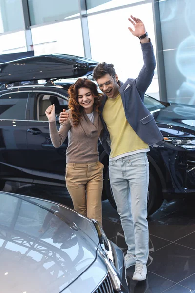 Homme heureux embrassant bouclé femme attrayante tout en célébrant triomphe dans la salle d'exposition de voiture — Photo de stock