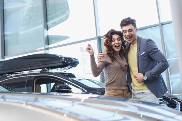 Homem alegre abraçando encaracolado mulher atraente enquanto celebra triunfo no showroom carro — Fotografia de Stock