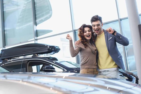 Homem feliz em óculos abraçando encaracolado mulher atraente enquanto celebra triunfo no showroom carro — Fotografia de Stock