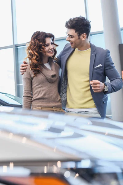 Messa a fuoco selettiva di uomo allegro in occhiali abbracciando bella donna riccia in showroom auto — Foto stock