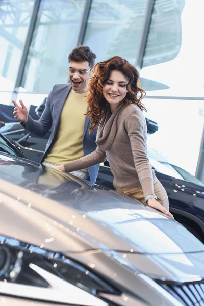 Селективное внимание веселой кудрявой женщины, улыбающейся рядом с взволнованным мужчиной в очках, смотрящим на автомобиль — стоковое фото