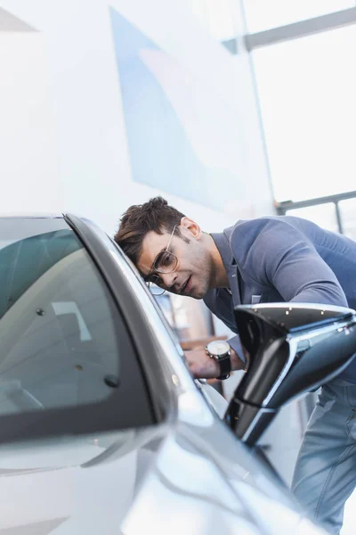 Selective focus of successful stylish man in glasses standing near auto in car showroom — Stock Photo