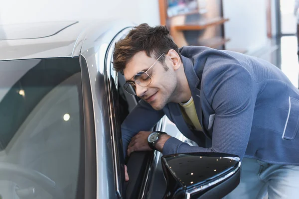 Foco seletivo de homem elegante alegre em óculos sorrindo perto de auto no showroom do carro — Fotografia de Stock