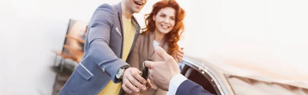 Colpo panoramico di rivenditore di auto dando le chiavi a uomo e donna allegro in auto showroom — Foto stock