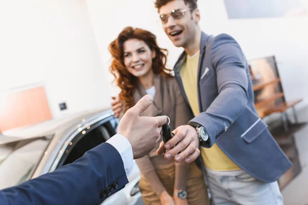 Vista ritagliata di rivenditore di auto dando le chiavi per l'uomo allegro e la donna in auto showroom — Foto stock