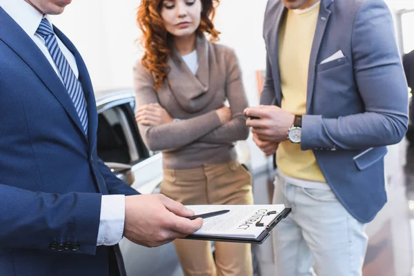 Vista ritagliata del rivenditore di auto in possesso di appunti con contratto vicino uomo e donna con braccia incrociate — Foto stock