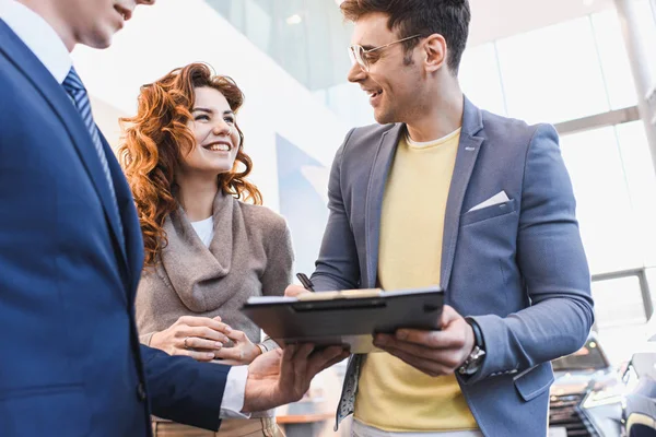 Cropped view of car dealer standing with cheerful man holding clipboard and looking at woman — Stock Photo