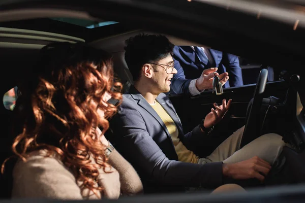 Vista recortada de concesionario de coches con llaves cerca de hombre alegre y mujer rizada en el coche — Stock Photo